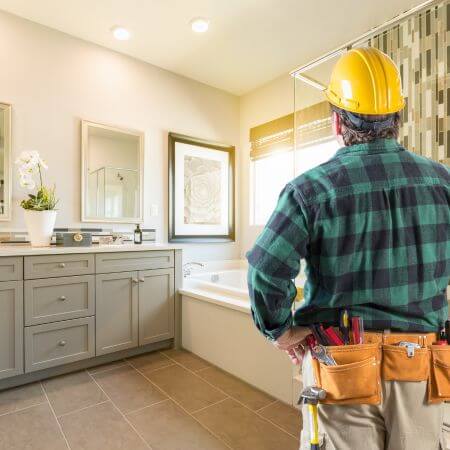 Bathroom Remodeling Company A man in a hard hat and plaid shirt stands in a bathroom, overseeing a remodeling project.