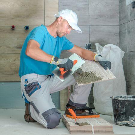 Professional-Bathroom-Designer A man kneels on a bathroom floor, carefully laying tiles as he works on the renovation project.